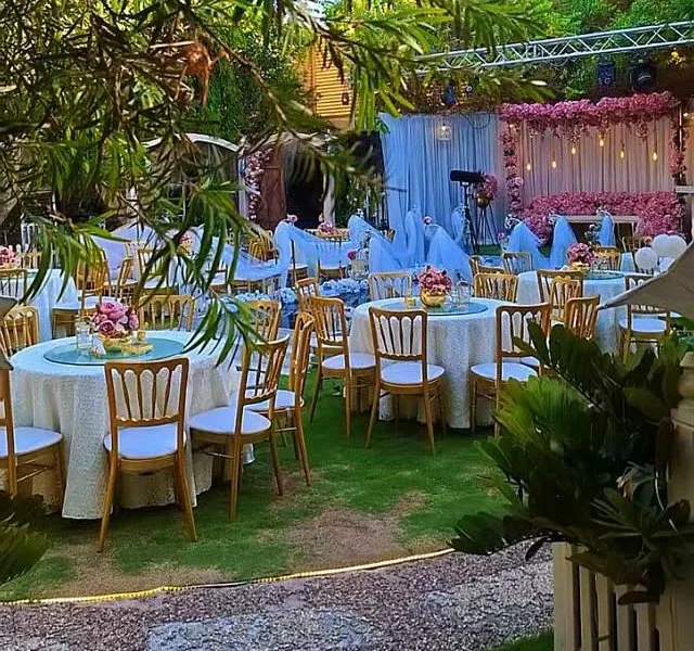 Round tables with lace cloths and gold chairs on a shaded lawn, with a pink floral stage behind